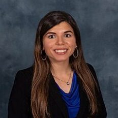 Professional headshot of a woman with long hair wearing a black blazer and blue blouse, smiling against a neutral background.