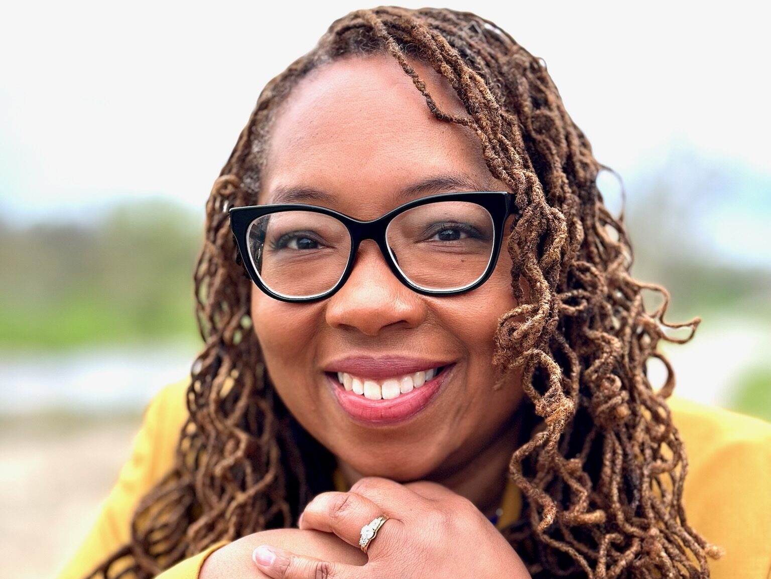 A woman with glasses and curly hair smiling while resting her chin on her hands.
