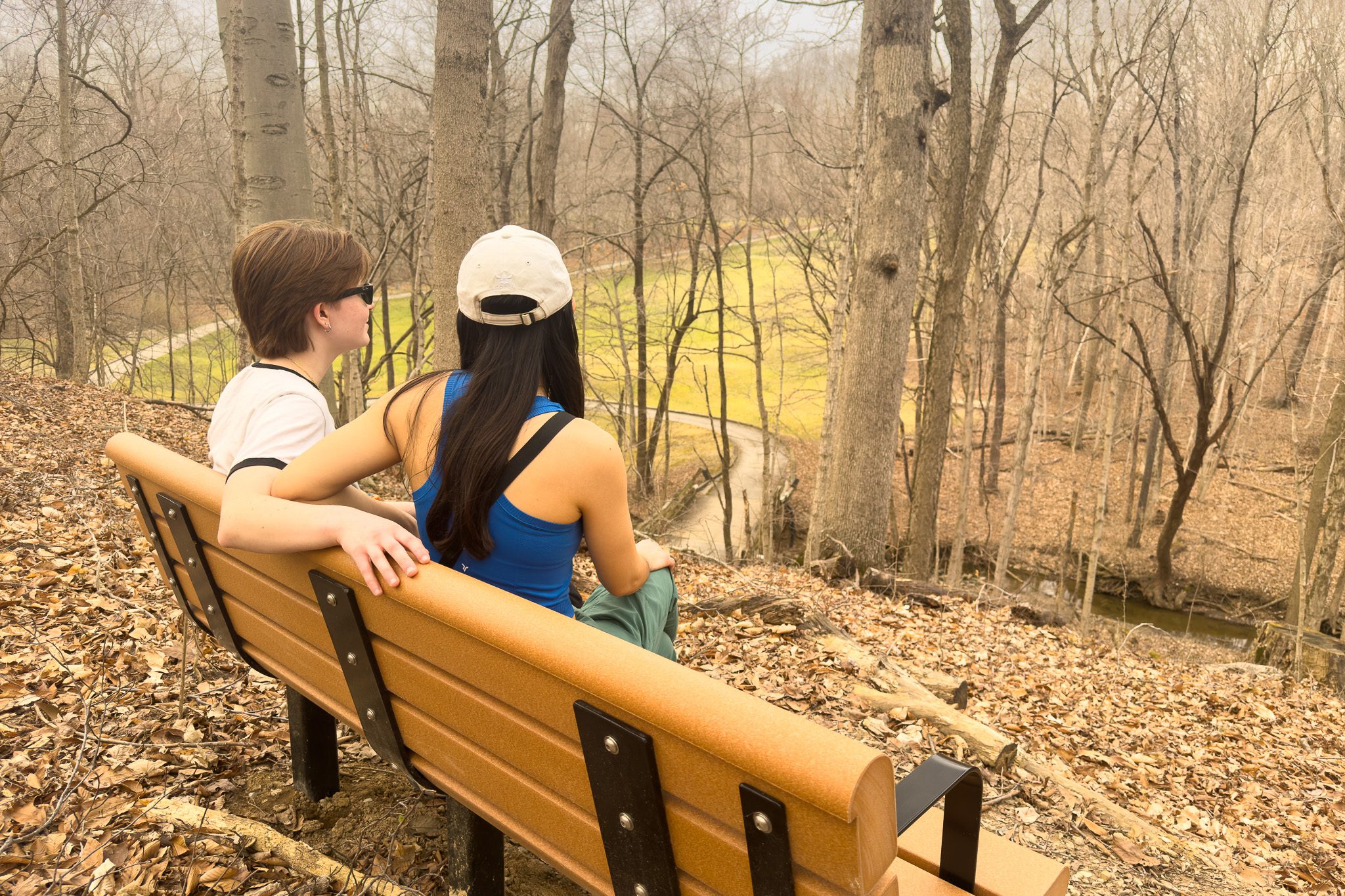 Two individuals seated on a bench in a wooded area, overlooking a trail and trees.