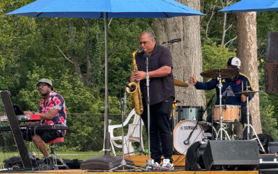 Musicians performing live at an outdoor concert with a saxophonist, keyboardist, and drummer.