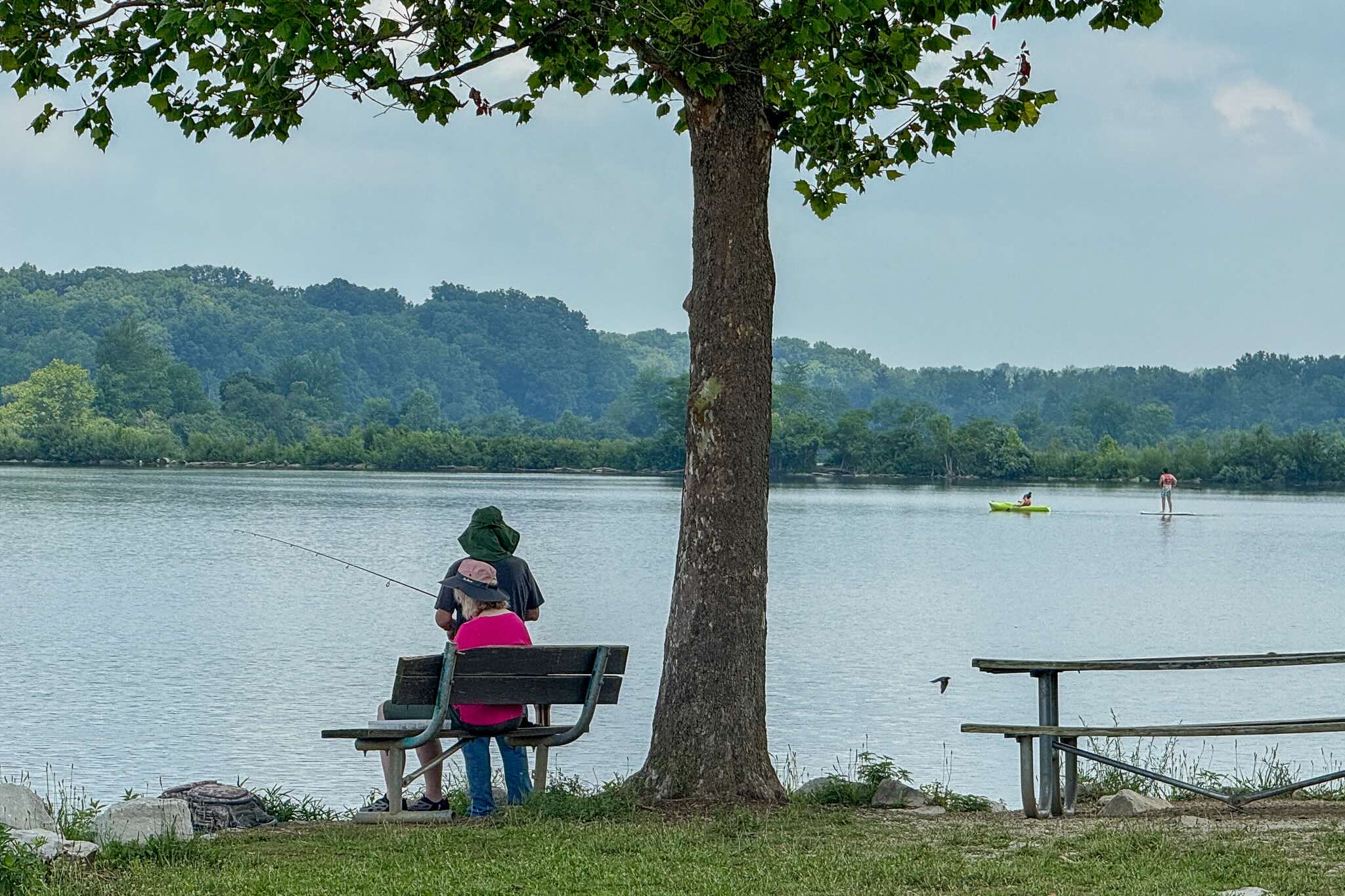 Two individuals fishing from a bench near a calm body of water with trees in the background.